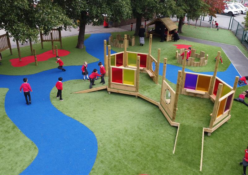 A playground with a wooden structure with two ramps either side. The structure has different colours of plastic sheets, creating a sensory area. The surface is artificial grass.